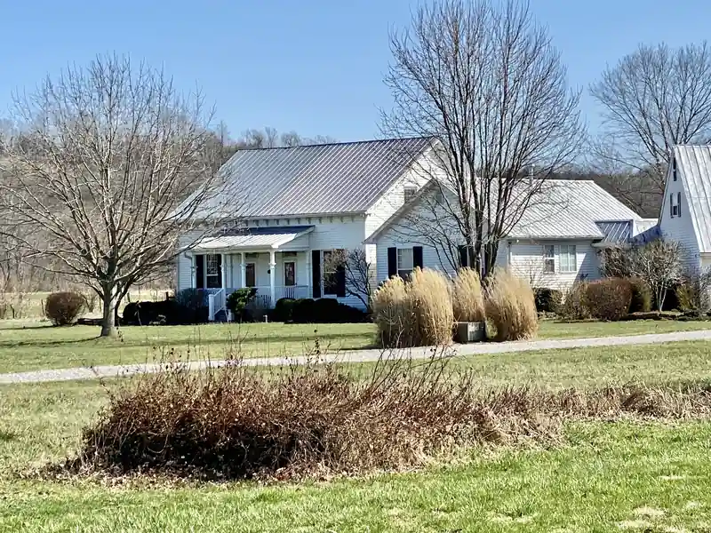 White farmhouse with a metal roof and a front porch, set back across a field with bare trees.