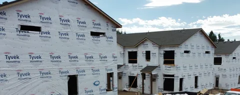 Two-story townhomes wrapped in house wrap, window frames cut in, on a new construction site.