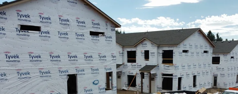 Two-story townhomes wrapped in house wrap, window frames cut in, on a new construction site.