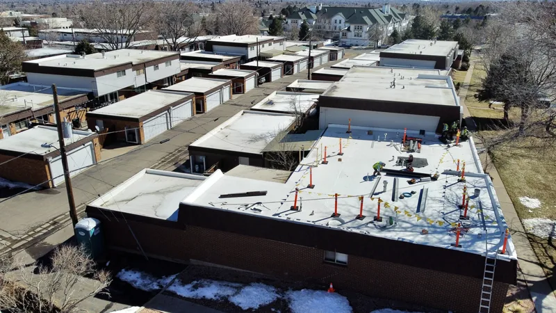White low-slope roof on a row of low brick buildings, with red safety flags and a ladder, from above.