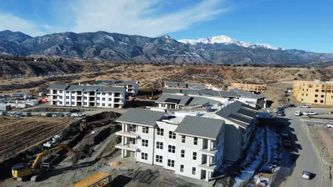 Apartment complex under construction with gray shingle roofs in place and excavators working on the site.