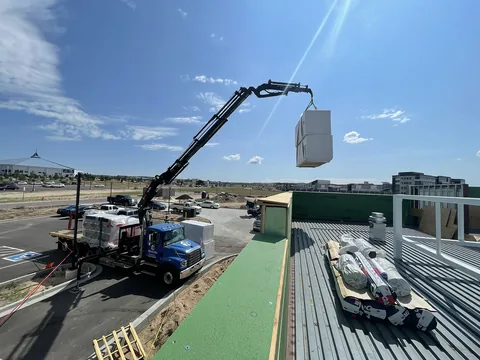 Boom truck lifting a pallet of roofing material up to a commercial metal-deck roof under a blue sky.