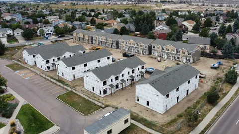 Townhome project under construction seen from above, house-wrapped buildings with gray shingle roofs and finished tan townhomes beyond.