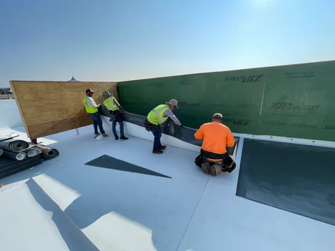 Four workers in high-visibility vests installing white TPO membrane against a green sheathed parapet wall.