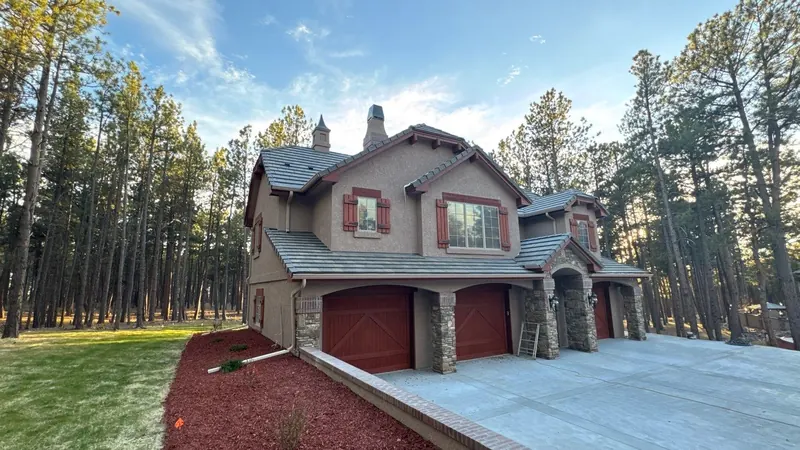 Stucco house with a gray flat concrete tile roof, red garage doors and a pine forest behind.