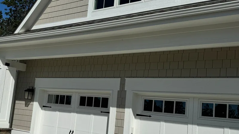 White gutter and eave over tan shake siding and two white carriage-style garage doors.