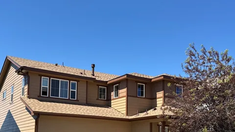 Tan two-story house with a light brown shingle roof, dark trim and a flowering tree beside it.