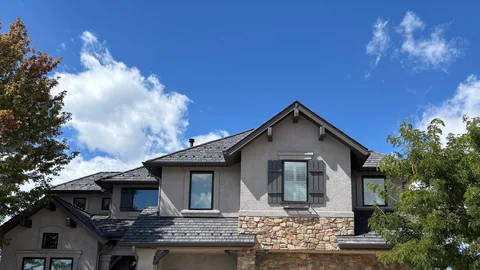 Stucco and stone house with a dark synthetic shake roof under a blue sky with white clouds.