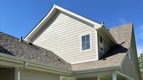 Tan lap siding gable and dormer above a brown shingle roof with roof vents and white gutters.