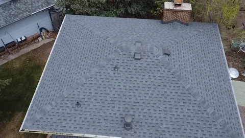 Overhead view of a gray shingle hip roof with ridge caps, roof vents and a brick chimney.