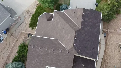 Overhead view of a brown shingle roof with several hips, valleys and vents above a gravel yard.