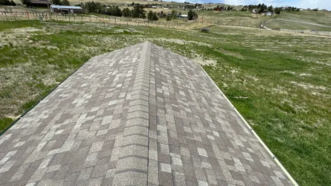 Hip ridge of a tan and gray shingle roof above a wide grassy field and distant houses.