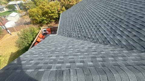 Gray architectural shingle roof planes meeting at a hip above a backyard deck with orange chairs.