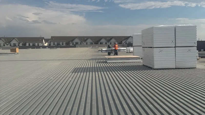 Wide low-slope metal roof deck with stacked insulation boards and two workers in the distance.