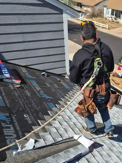 Roofer in a safety harness and tool belt laying gray shingles over synthetic underlayment.