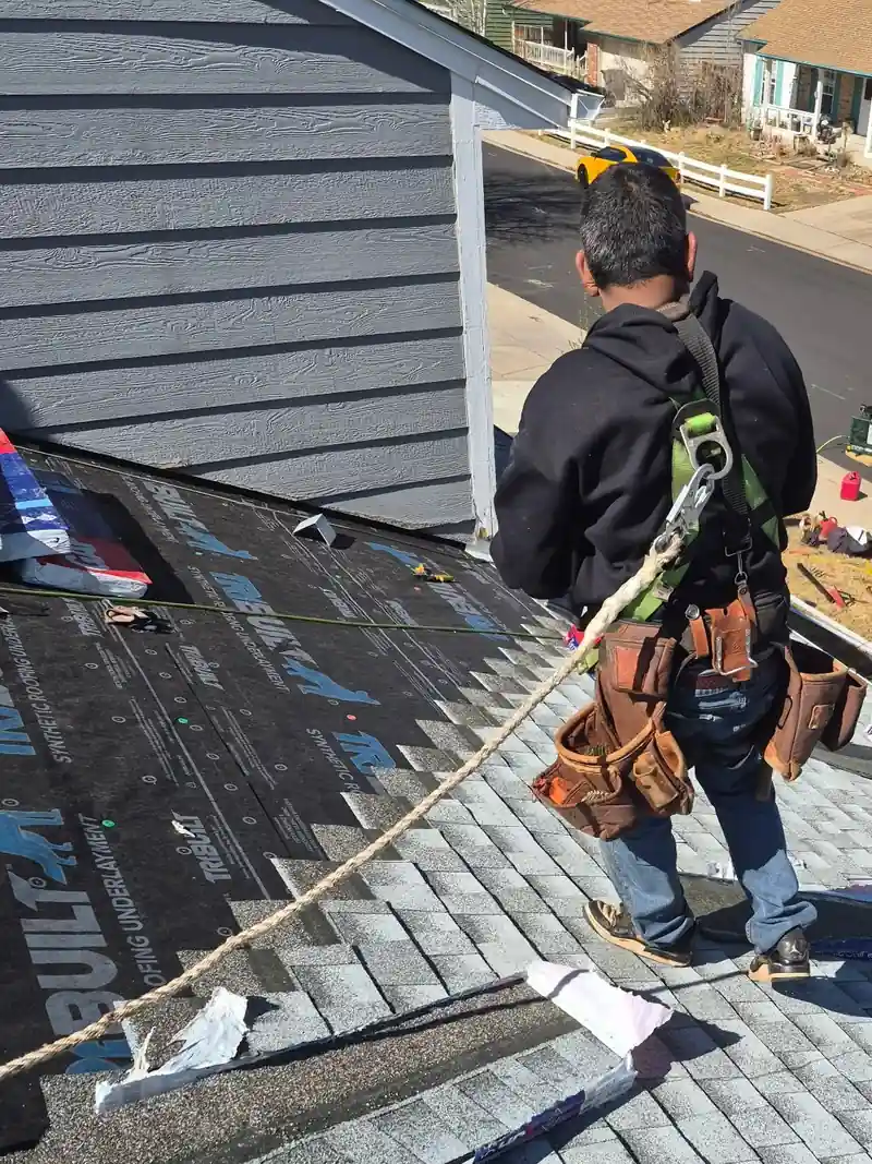Roofer in a safety harness and tool belt laying gray shingles over synthetic underlayment.