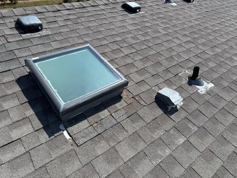 Aged skylight with sealant smears, a bent metal vent and a pipe boot on weathered gray shingles.