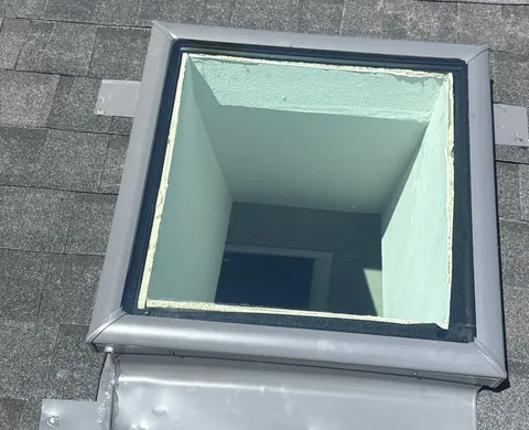 Straight-down view into a skylight shaft with a gray metal frame and flashing on dark shingles.