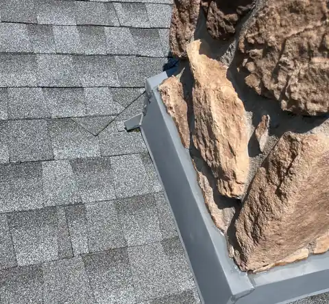 Gray metal flashing wrapped around the corner of a stacked stone chimney on gray shingles.