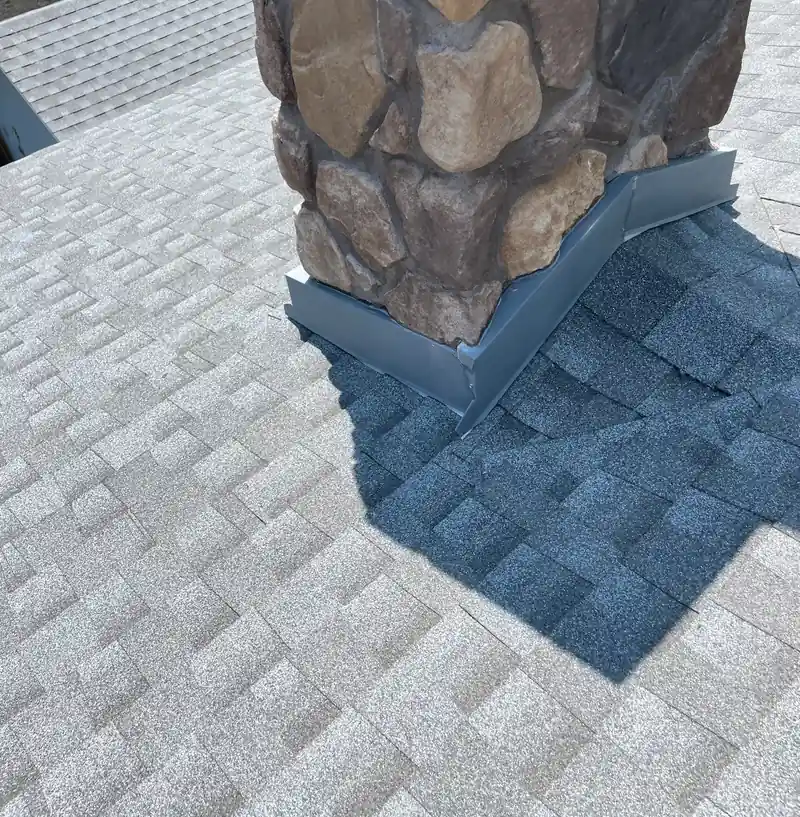 Base of a stacked stone chimney with gray metal flashing on light gray shingles.