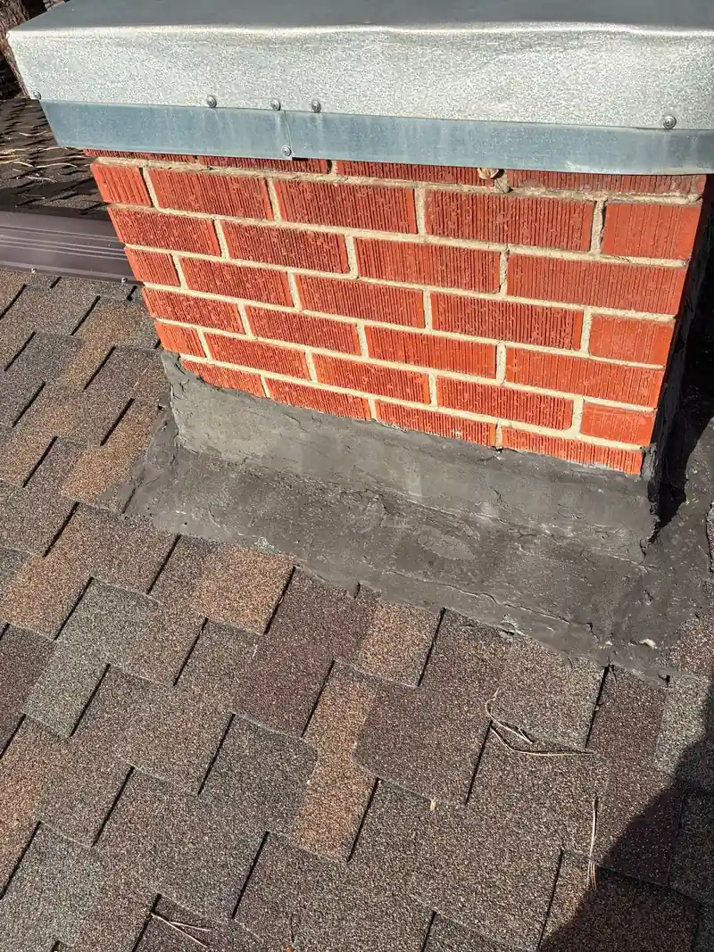 Red brick chimney with a metal cap and a thick roof cement base on brown shingles.