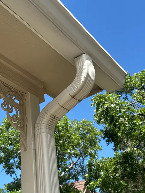 Beige downspout with a creased elbow under a wide eave beside a carved porch bracket and green trees.