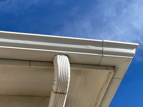 White gutter corner with a split seam and a cracked downspout under a stained soffit.