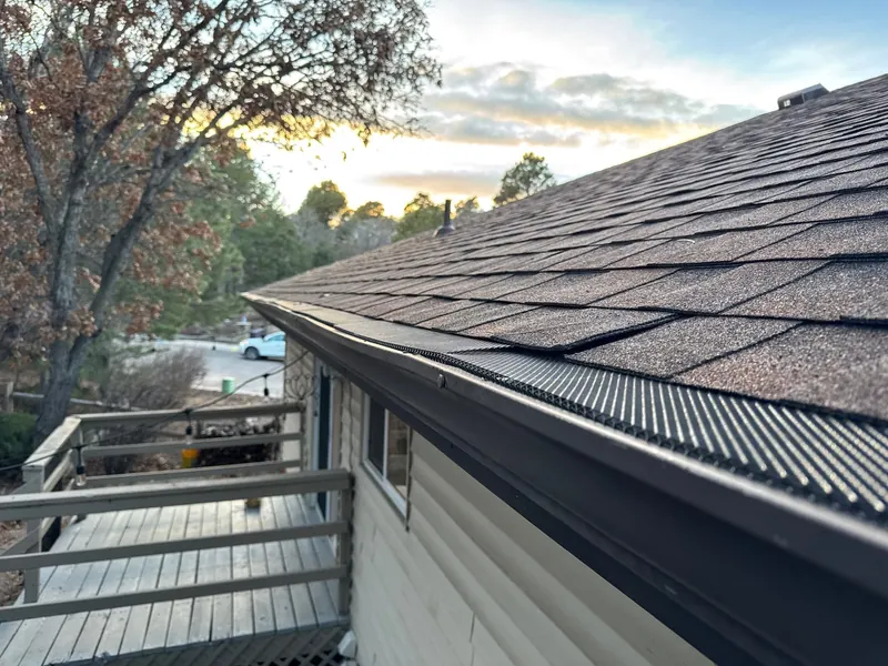 Black gutter guard along a brown shingle roof edge beside a wooden deck at dusk.