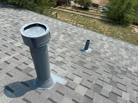 Gray painted vent stack and a smaller pipe boot on a light gray shingle roof above a yard.