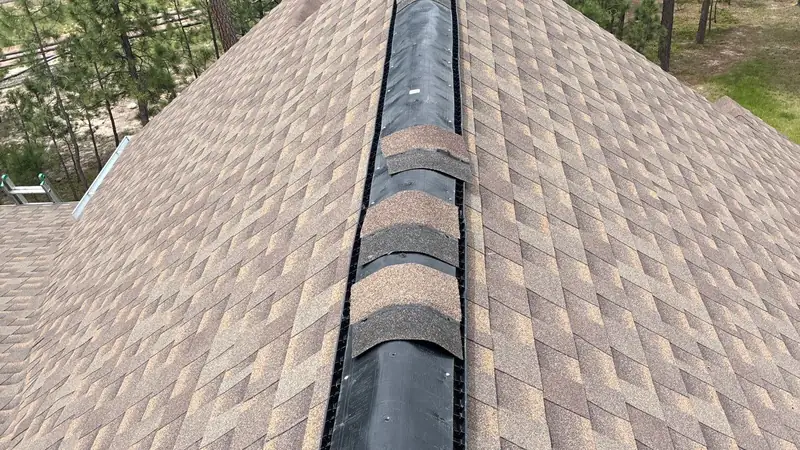 Black ridge vent partly covered with tan cap shingles along the ridge of a brown shingle roof.